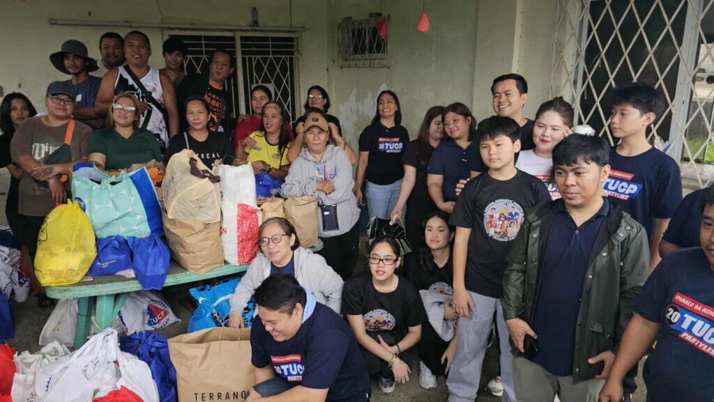 A group of people pose with bags of supplies, many wearing "TUCP Partylist" shirts.